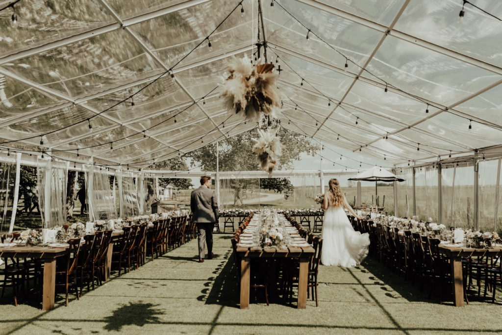 bride and groom walk through their wedding reception space which is a clear tent with hanging pampas grasses, long harvest tables adorned with rows of romantic florals. www.thegathered.ca