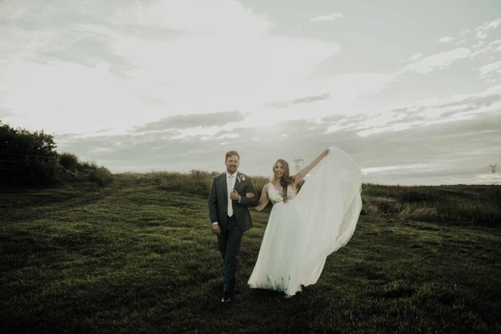bride holds her dress up in one hand as she walks arm in arm with the groom down a grassy hill in a prairie field. www.thegathered.ca