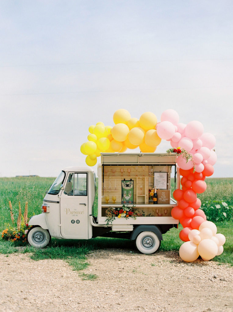 yellow, pink and red balloons surround a small Prosecco cart while bright blue skies and green field act as a backdrop www.thegathered.ca