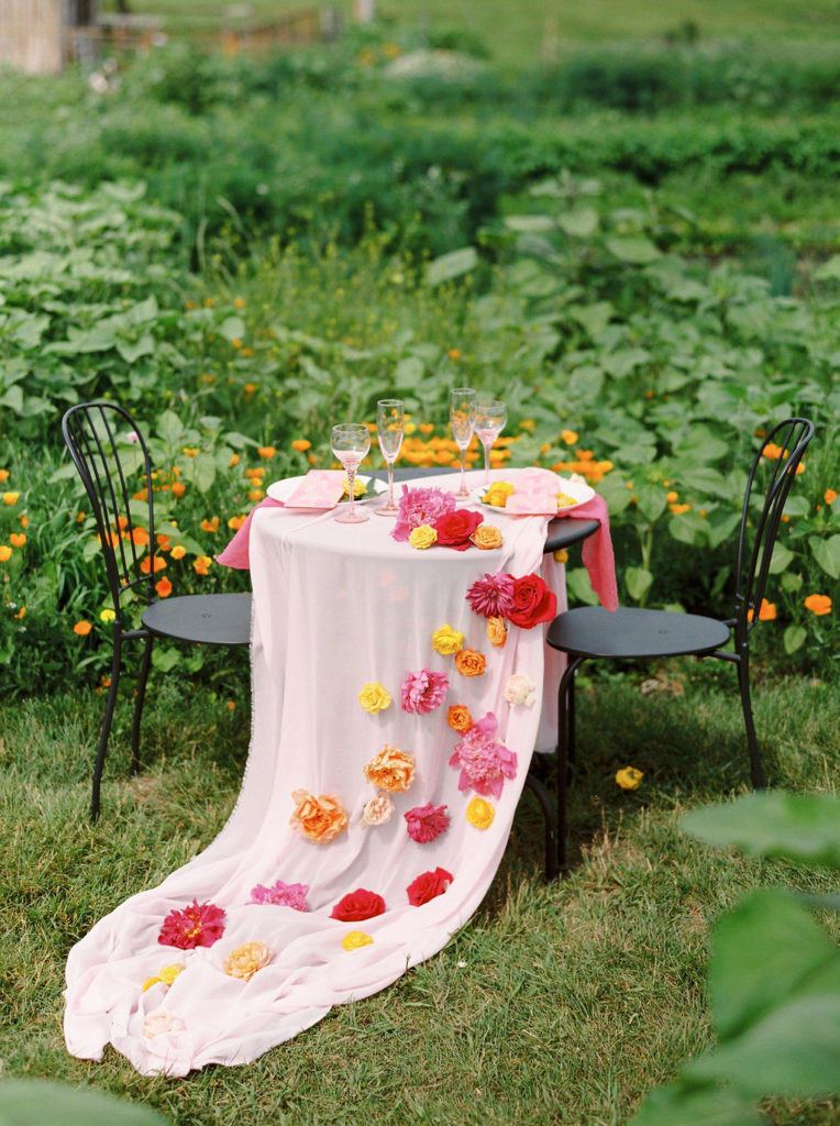 bistro table placed in a green garden with blush pink tablecloth and bright floral accents