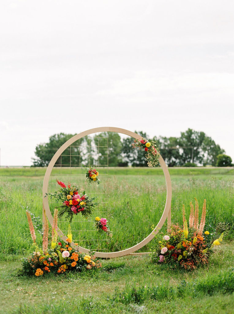 circular arbor decorated with bright pink, orange and yellow flowers while a green prairie field act as the backdrop