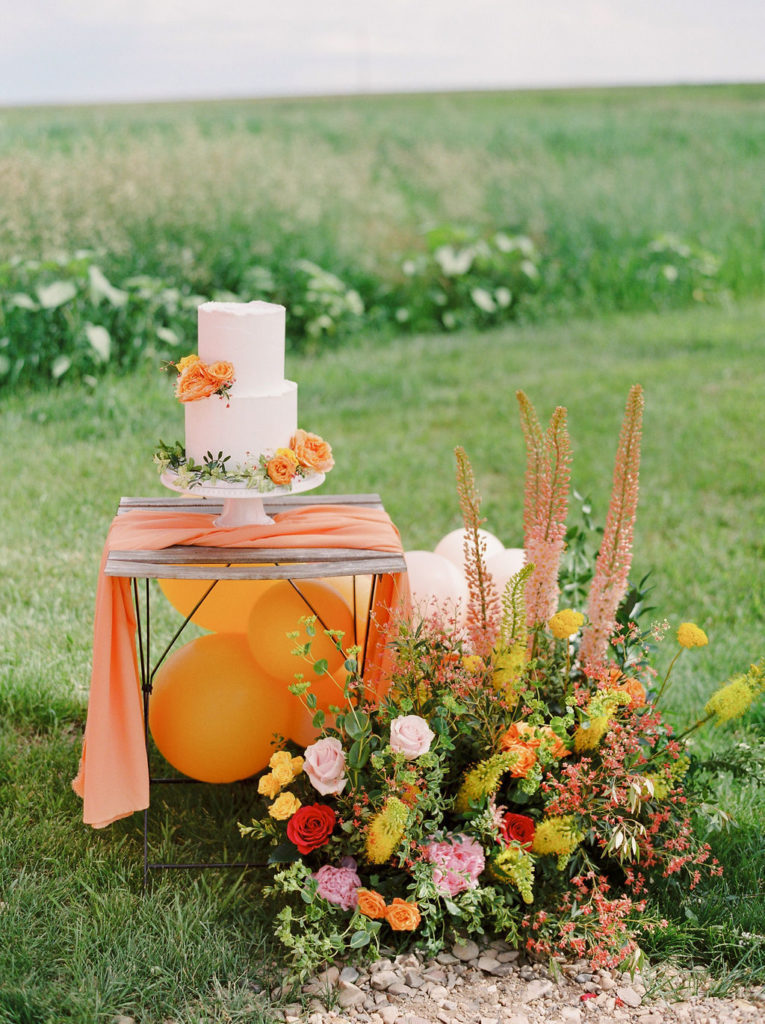 bistro table with orange table linens a white cake and bright colored floral arrangement in a grassy green field. www.thegathered.ca