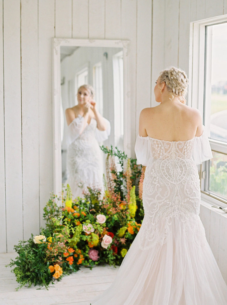 white cottage with a bride in a lacy dress standing in front of a mirror, bright green, orange and pink floral arrangements adorn the floor making a pop against the white walls. www.thegathered.ca