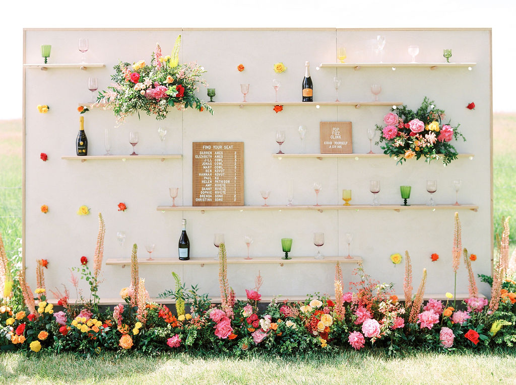 wood letter board lists the names of the guests on a display wall filled with wine goblets and bright florals