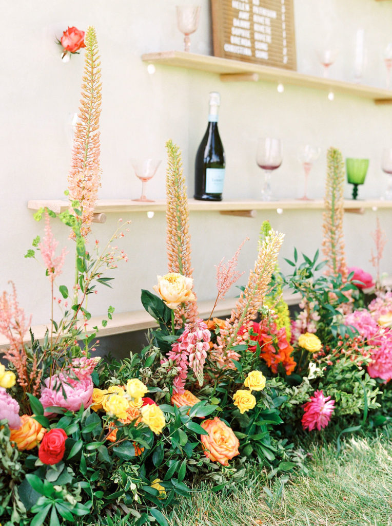 display wall filled with colored wine goblets, seating charts and bright pink and orange floral arrangements.