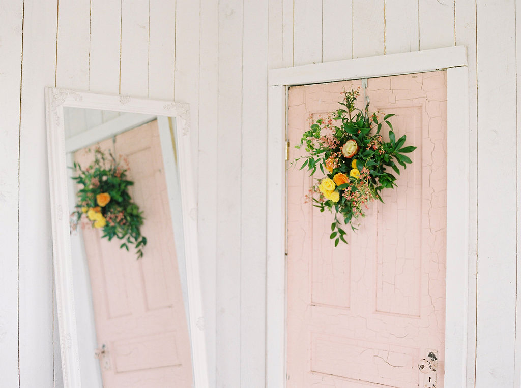 blush pink antique door, hanging on the door is a bright green, yellow and orange flower arrangement acting as the perfect spring setting for this shoot