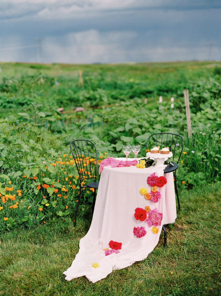 bistro table with pink table cloths and bright red and pink flowers with bright green garden in the background.