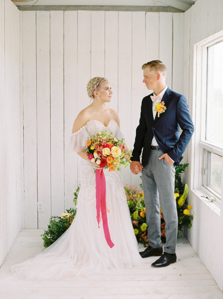 couple in a white cottage makes the perfect backdrop for the brides bright pink, orange and yellow flower bouquet. www.thegathered.ca