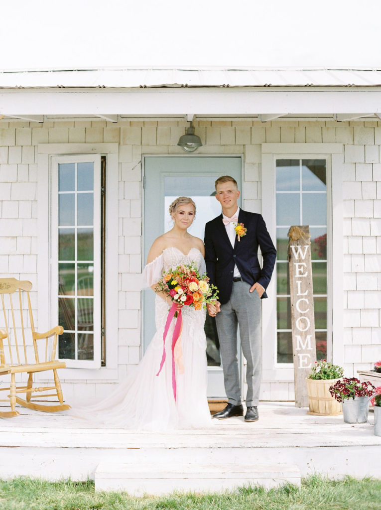 white cottage with a rocking chair on porch and a couple with bold toned florals making a pop against their wedding attire.