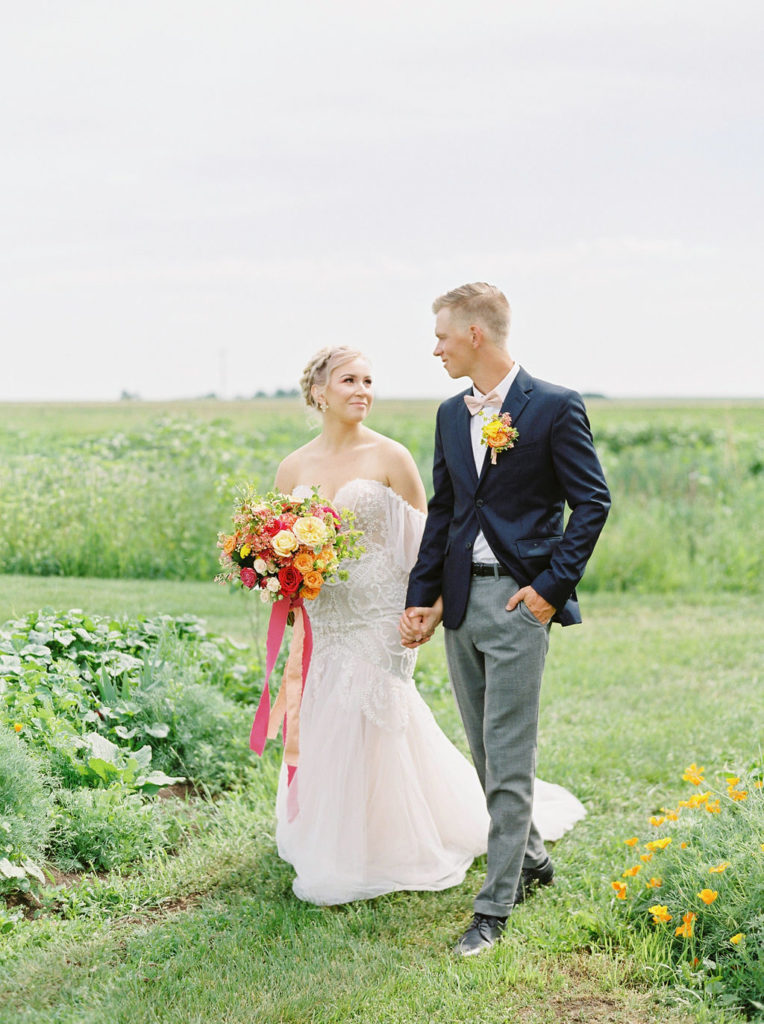 couple walks in a bright green spring field while the brides bouquet shines with bright red and orange flowers