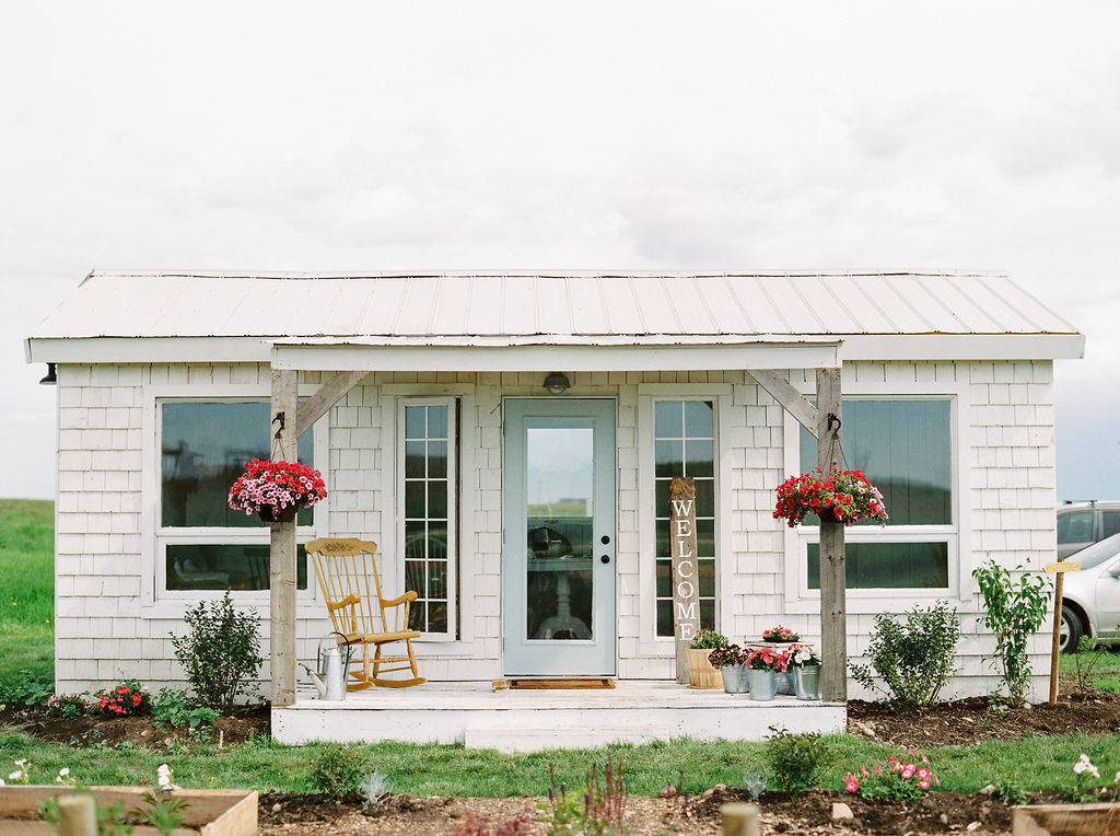 white cottage with blue door is surrounded by pink hanging flowers www.thegathered.ca