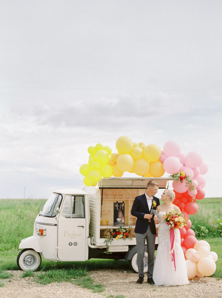 yellow, pink and red balloons surround a small Prosecco cart while a couple cheers in front.