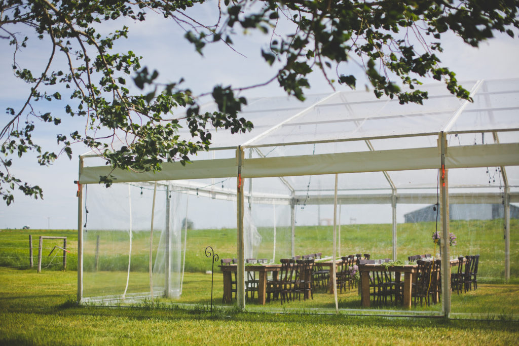 clear wedding tent in a prairie field with wooden tables and chairs at the gathered