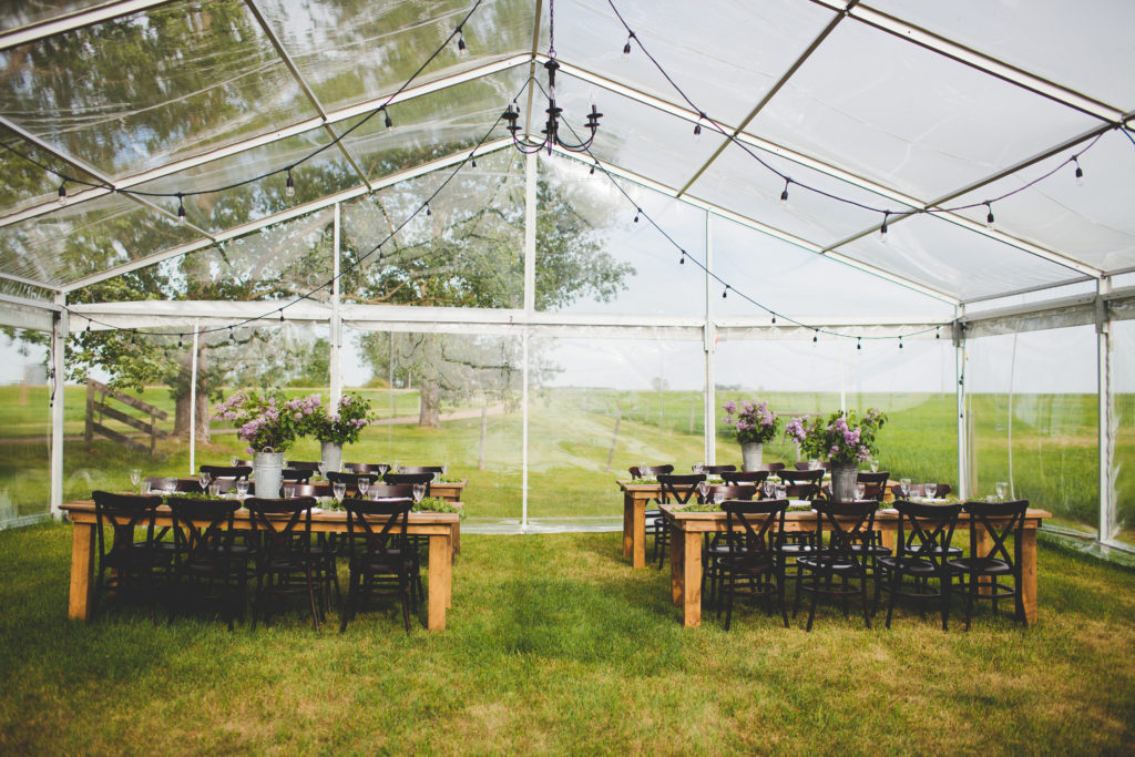 clear tent with hanging globe lights in this prairie setting. Wood harvest tables and chairs decorated with lilacs - the perfect spring decor. www.thegathered.ca