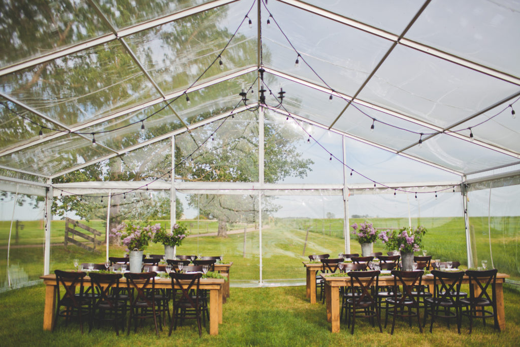 clear tent with hanging globe lights in this prairie setting. Wood harvest tables and chairs line the tent with lilac floral center pieces . www.thegathered.ca