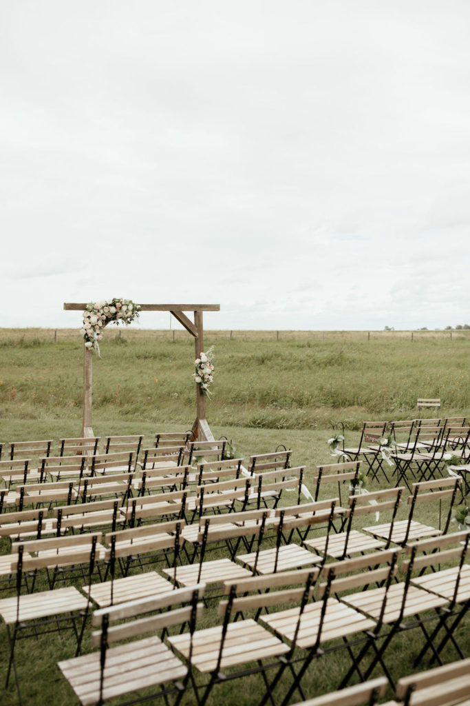 Prairie Ceremony space with wood chairs. Decorated with soft pinks and white flowers giving a muted appearance in the overcast sky in Alberta, Canada. The Gathered