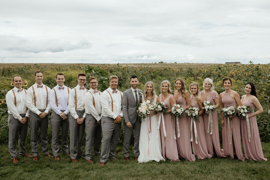 bridal party dresses in pink tones surround the bride and groom as they all stand in front of a flower garden. www.thegathered.ca