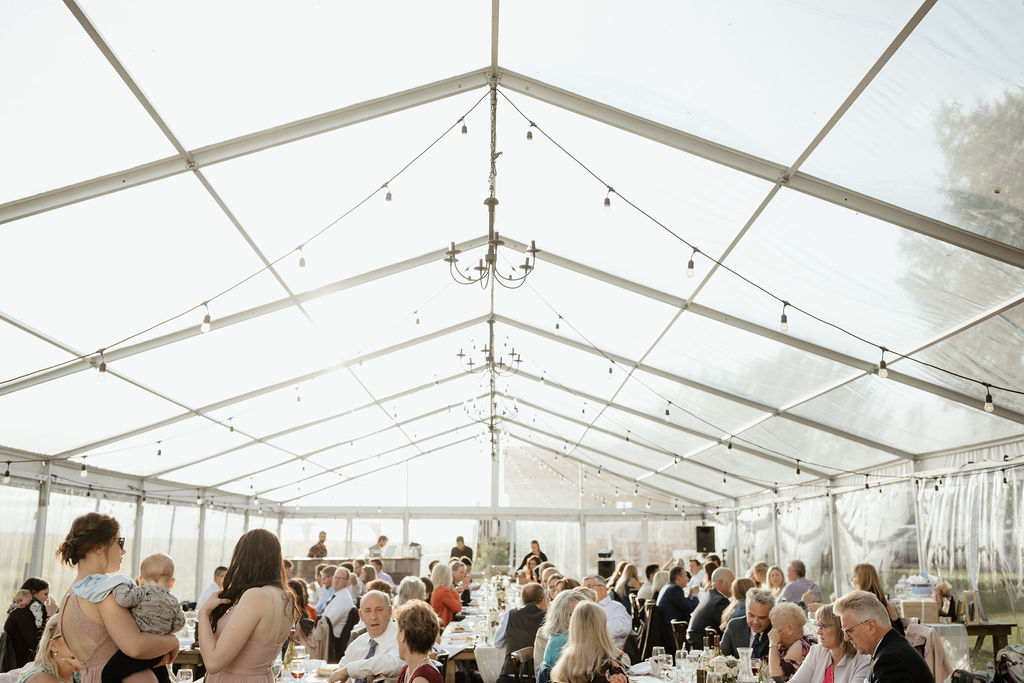 clear tent with hanging globe lights in this prairie setting. Wood harvest tables and chairs with guests mingling - the perfect setup. www.thegathered.ca