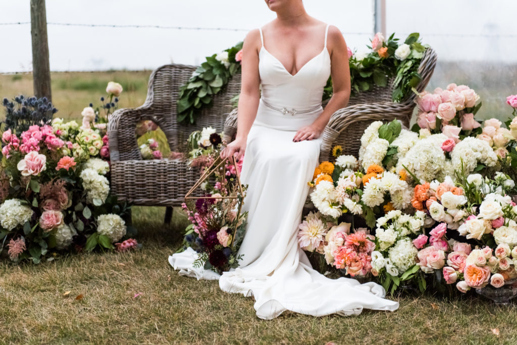 a bride poses on a wicker chairs completely surrounded by flowers, the tones are mainly white, pinks, peach