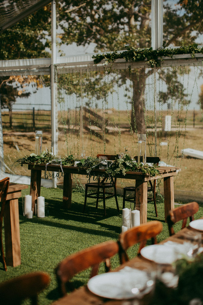 in a clear tent a sweetheart table is setup with layers of greenery running along the table, pieces of flowing greens drape down behind the table, the gathered