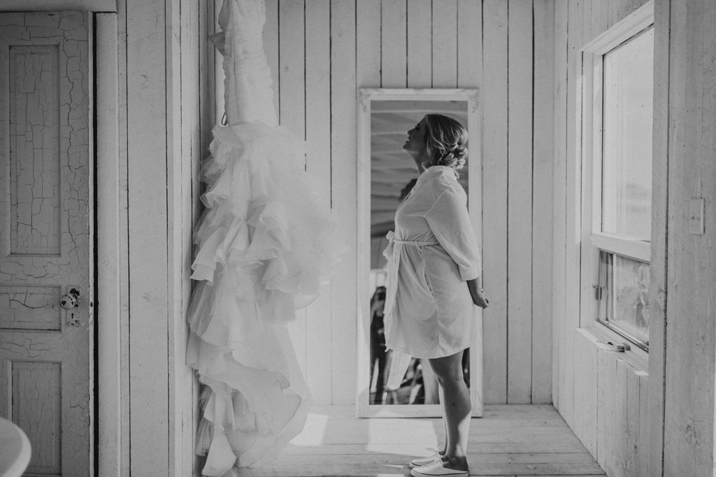 a black and white photo of a bride standing in a cottage looking up at her dress hanging on the wall with her hands behind her back