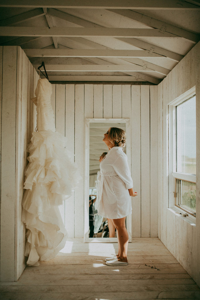 a bride standing in a cottage looking up at her dress hanging on the wall with her hands behind her back