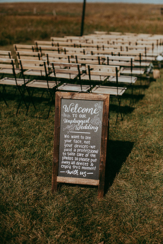 a prairie field ceremony space with wood slotted chairs and a sign that says "welcome to our unplugged wedding"