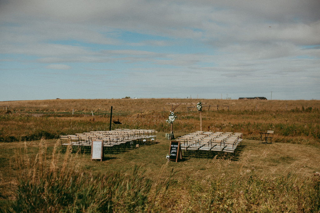 Prairie Ceremony space with wood chairs. Decorated with soft pinks and white flowers giving a muted appearance in the overcast sky in Alberta, Canada. The Gathered