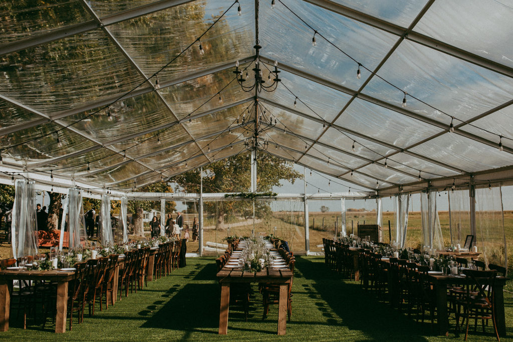 clear tent with hanging globe lights in this prairie setting. Wood harvest tables and chairs decorated with greenery and candles in hurricane vases. www.thegathered.ca
