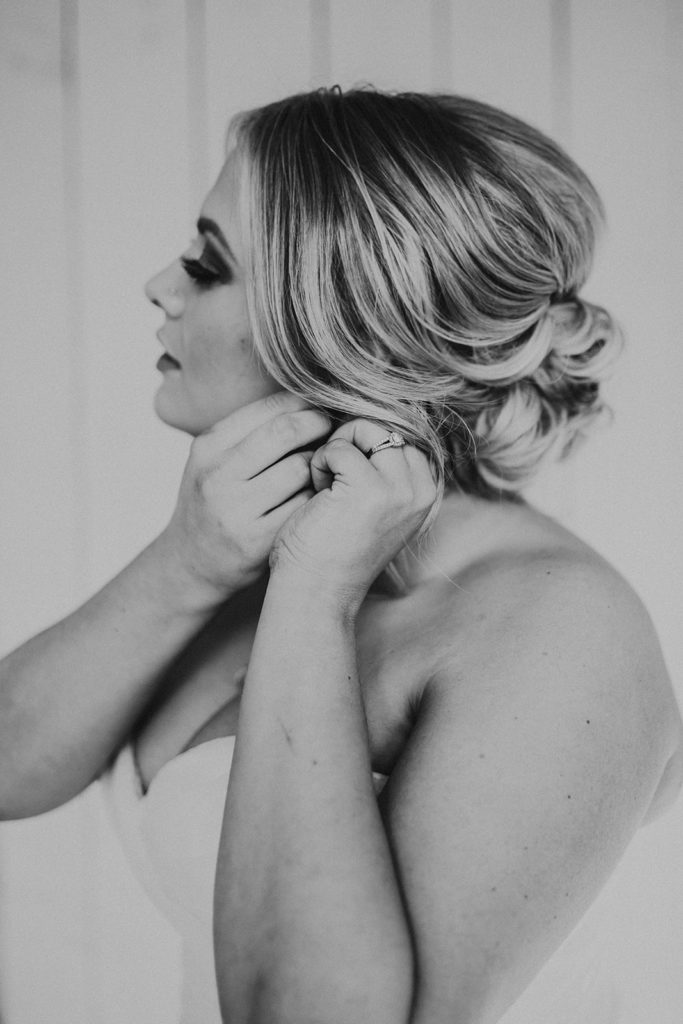 black and white photo of a bride putting on her earrings with her hair in a lowered loose bun