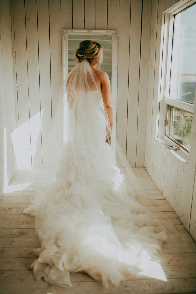 in a white cottage a bride with a flowing train and long veil stands beside the window where a warm fall sun streams in