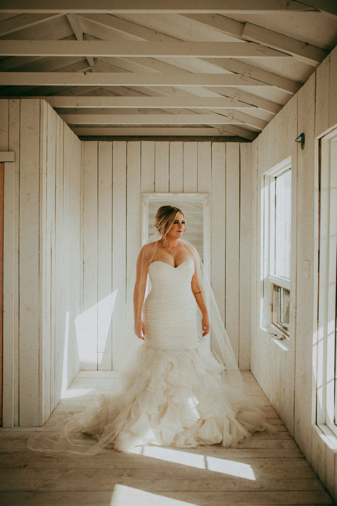 in a white cottage a bride with a flowing train and long veil stands beside the window where a warm fall sun streams in