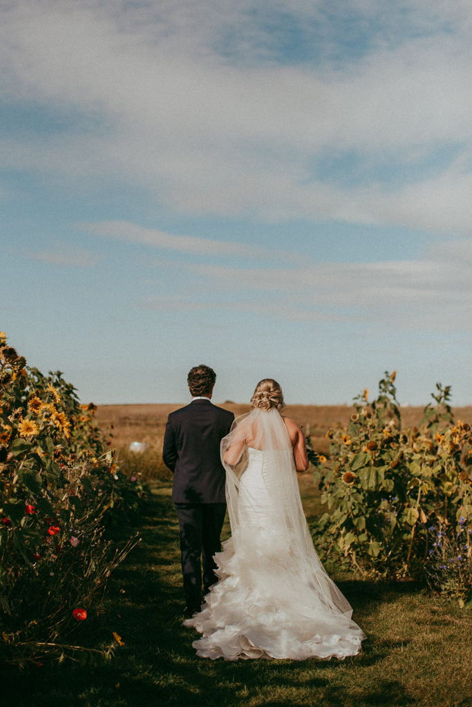 a bride and her father walk down a sunflower laden aisle with large trees in the background, the sun casts a warm fall glow over the whole scene