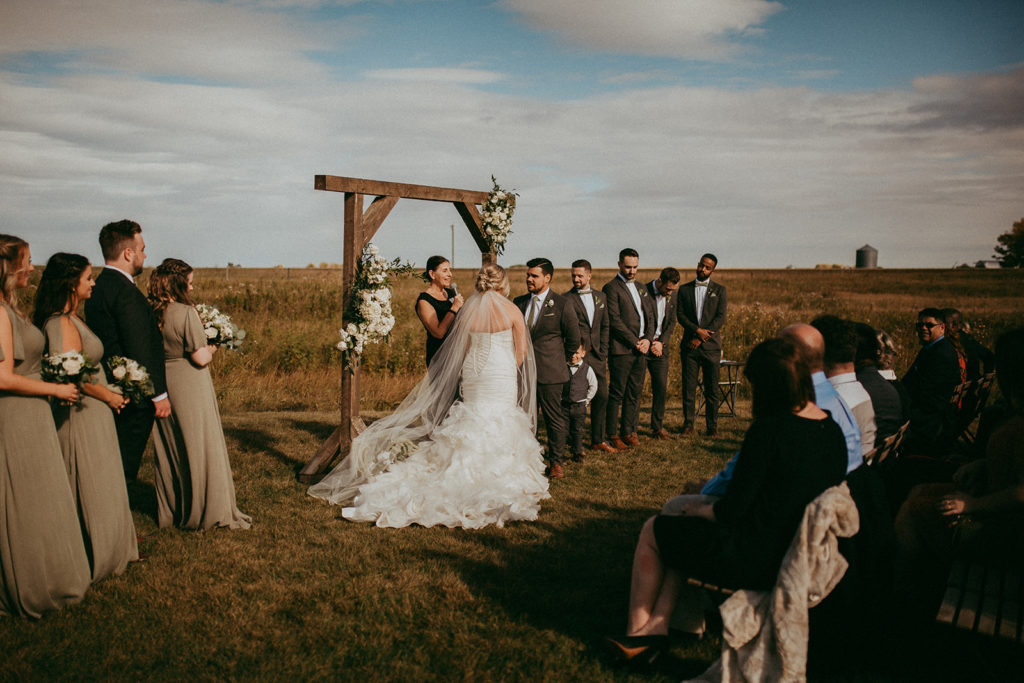 in a prairie ceremony space a large bridal party in shades of green and grey stand around a couple as they say their vows