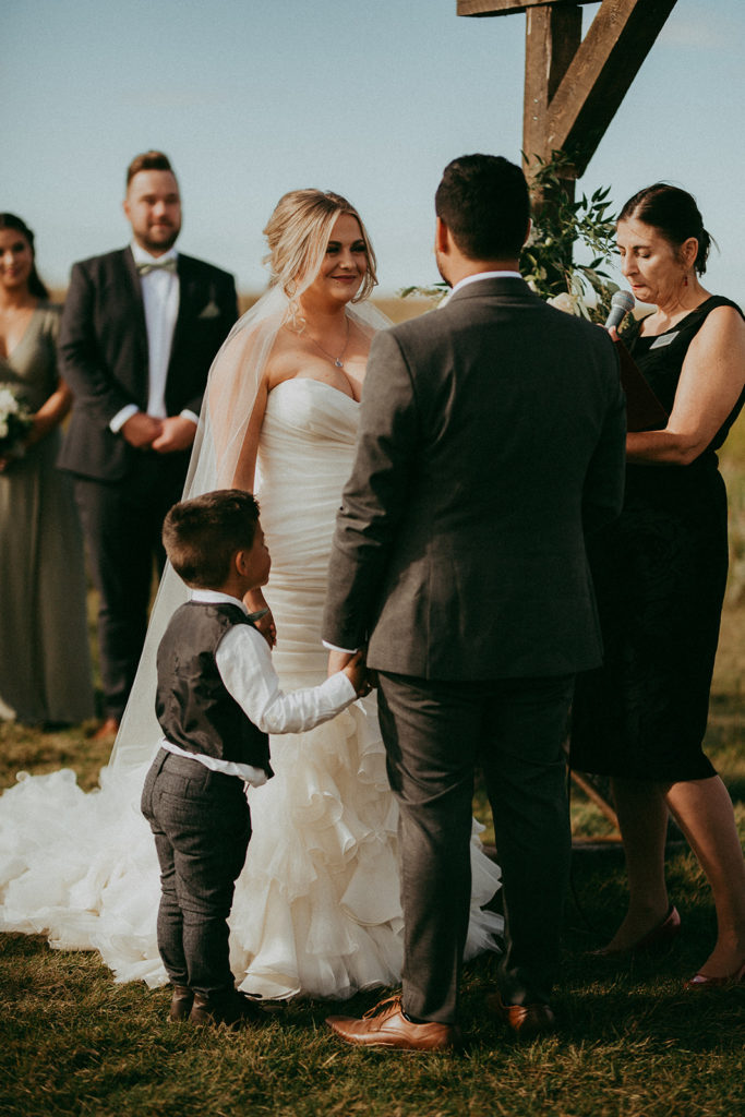 a bride and groom and their son hold hands as they say their weddings vows in a outdoor ceremony