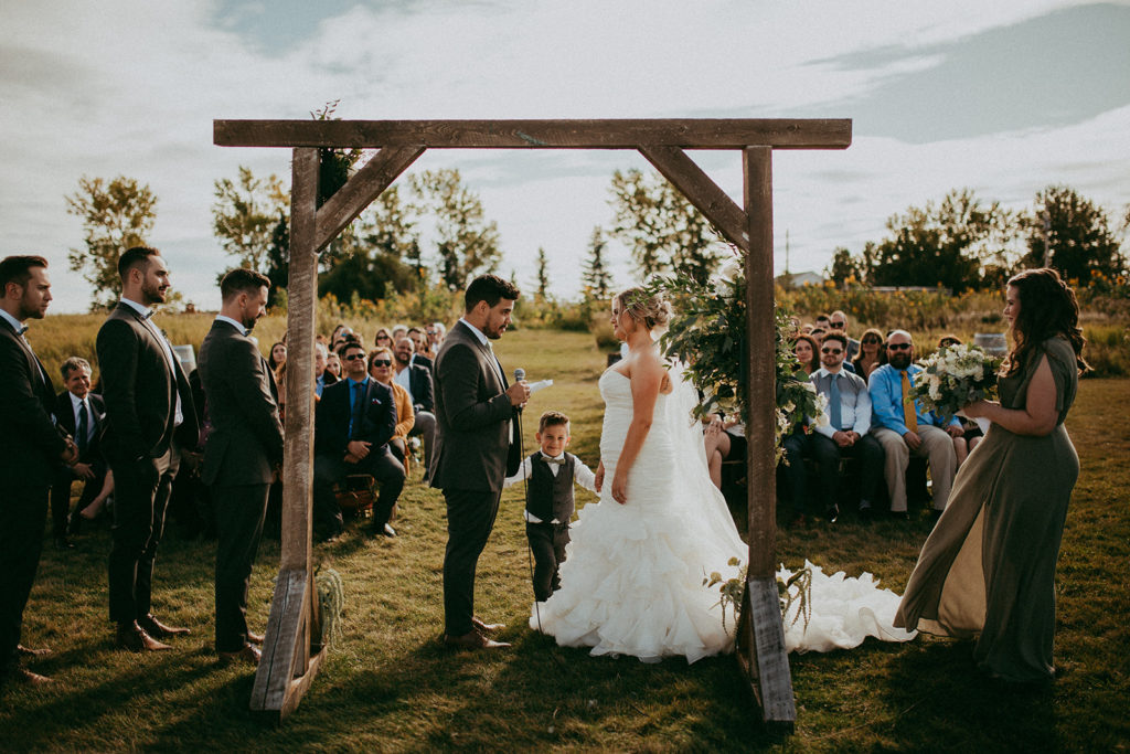 in a field weddings guests watch as a bride and groom and their son exchange vows under a wooden arbor - the gathered