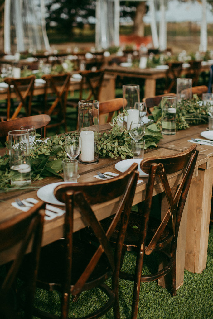 wooden harvest tables lined with greenery and pillar candles in hurricane vases