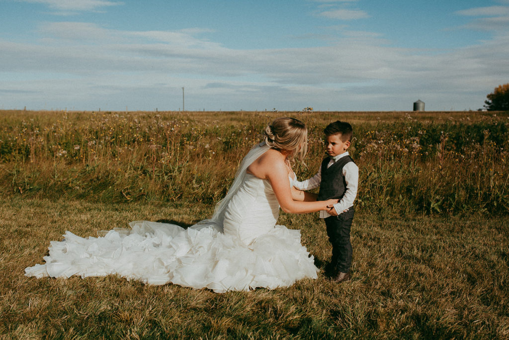 a bride kneels in a prairie field talking to her young son while the fall sun shines on them