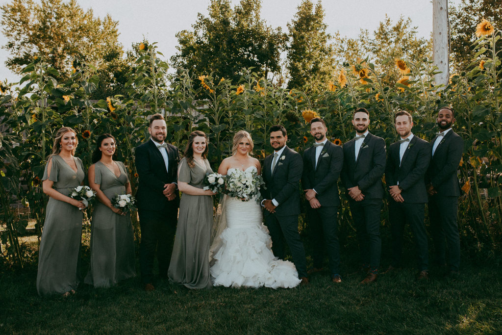 a wedding party in shades of green stand in front of large sunflowers that tower overhead, thegathered.ca