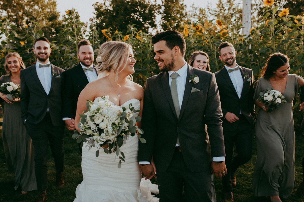 a bride and groom walk in front of their bridal party as the gaze at each other, large yellow sunflowers make an appearance behind them