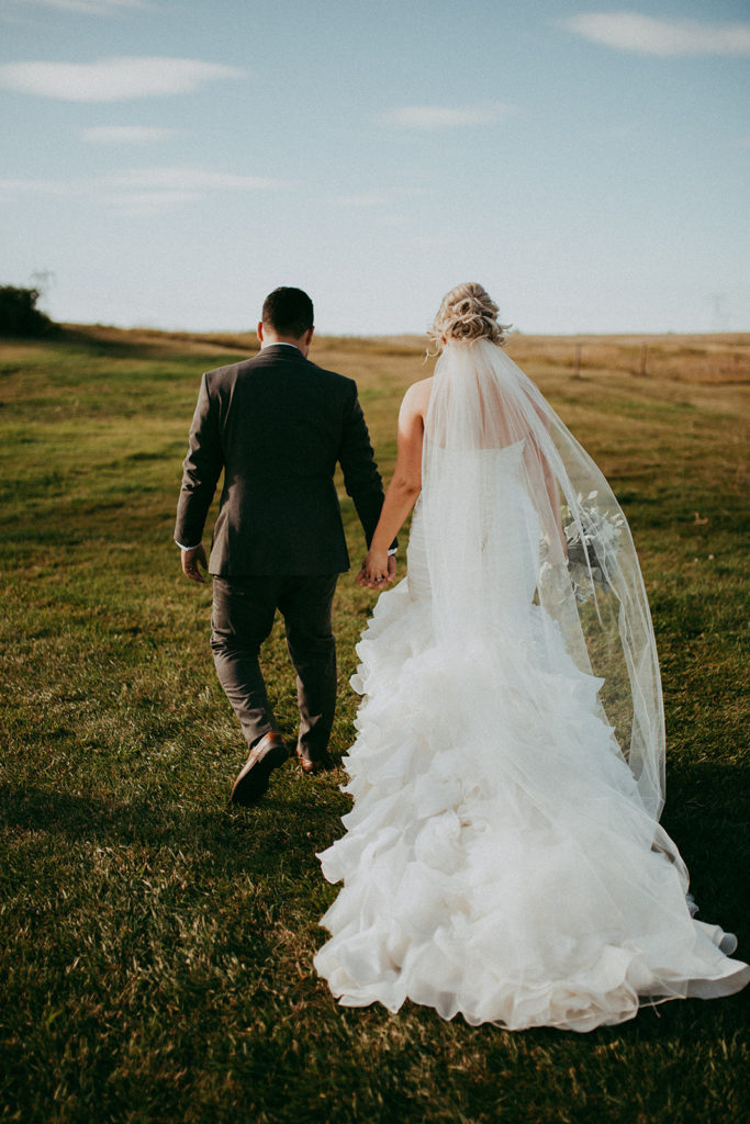 a bride and groom walk hand in hand into an open prairie field