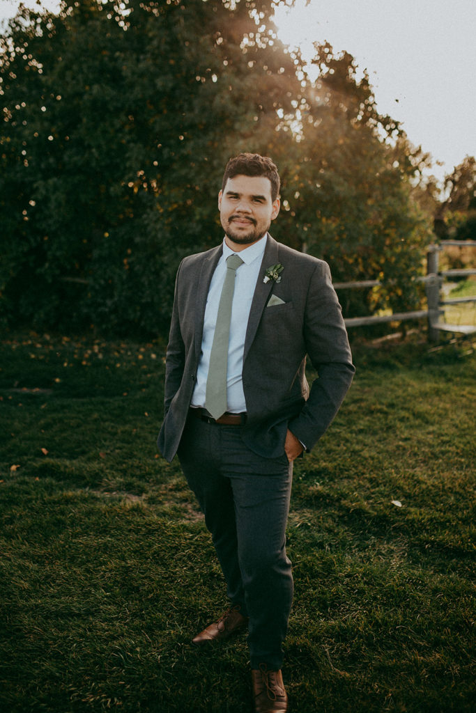 groom stands for a photo in a dark grey tux with a green tie and pocket square