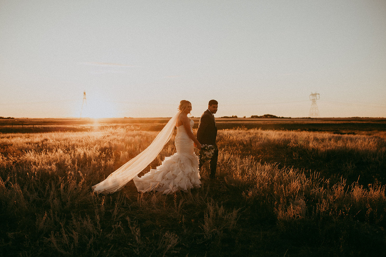 a bride and groom walk through an open prairie field while the sun sets behind them
