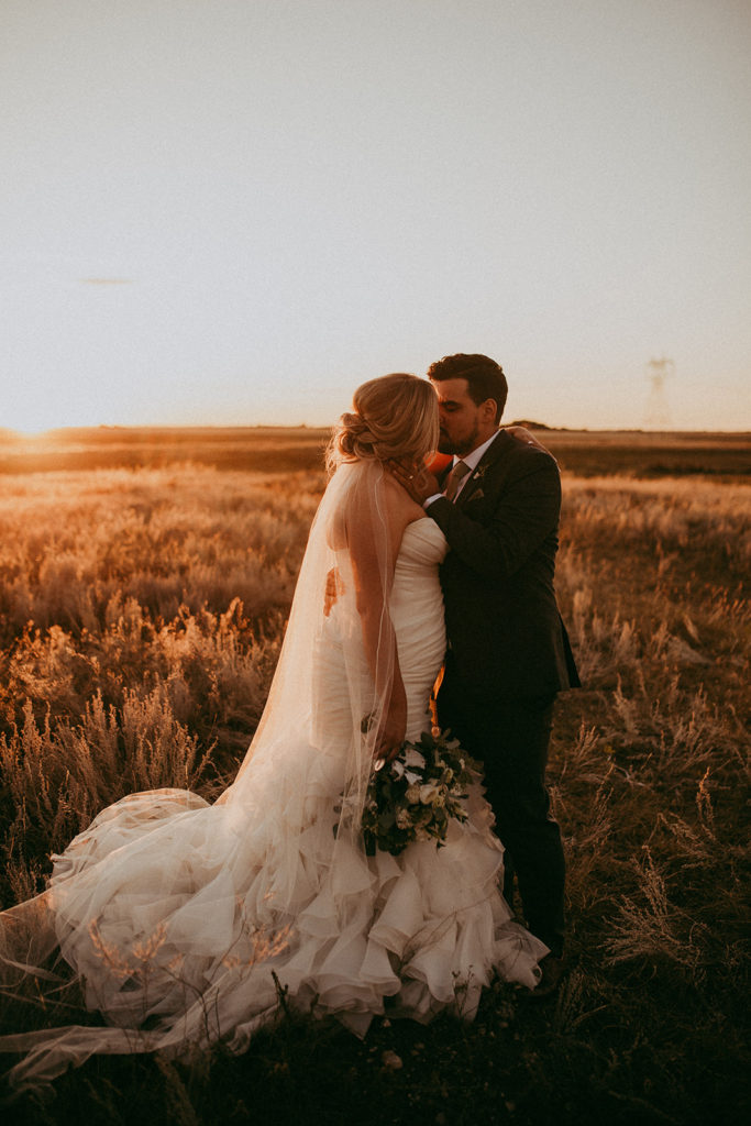 bride and groom kiss in a golden prairie field at sunset