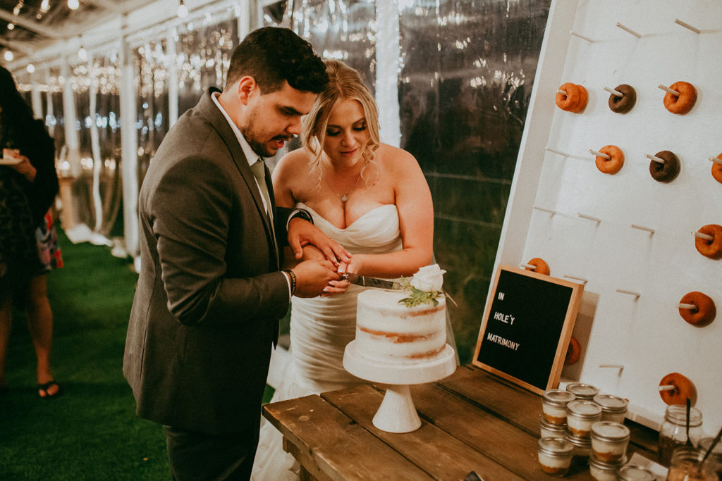 bride and groom cut their wedding cake in a clear tent reception space, a donut wall sits off to the side of them