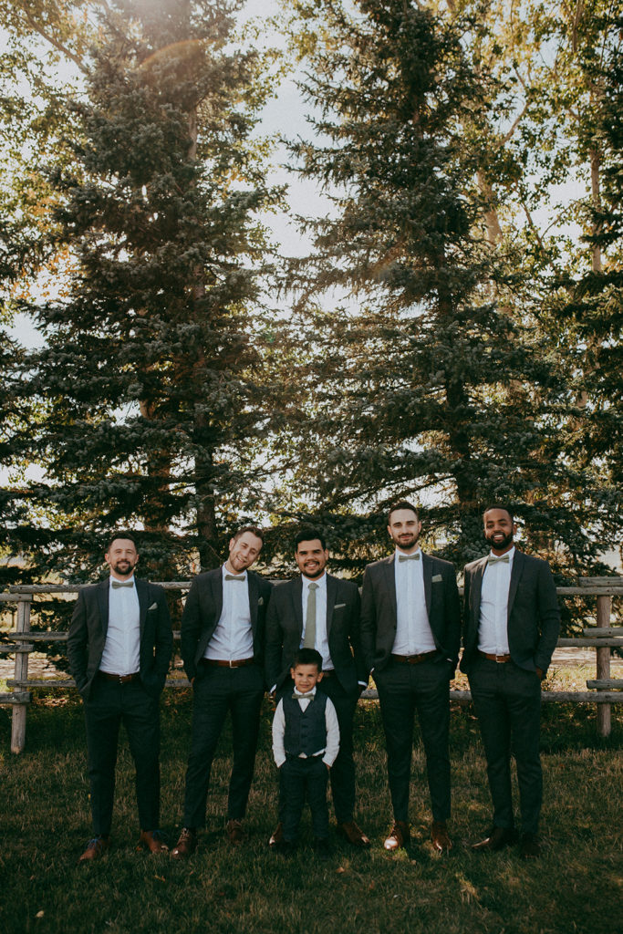 groomsmen and ring bearer stand against trees with the sun setting in the background in the beautiful prairies