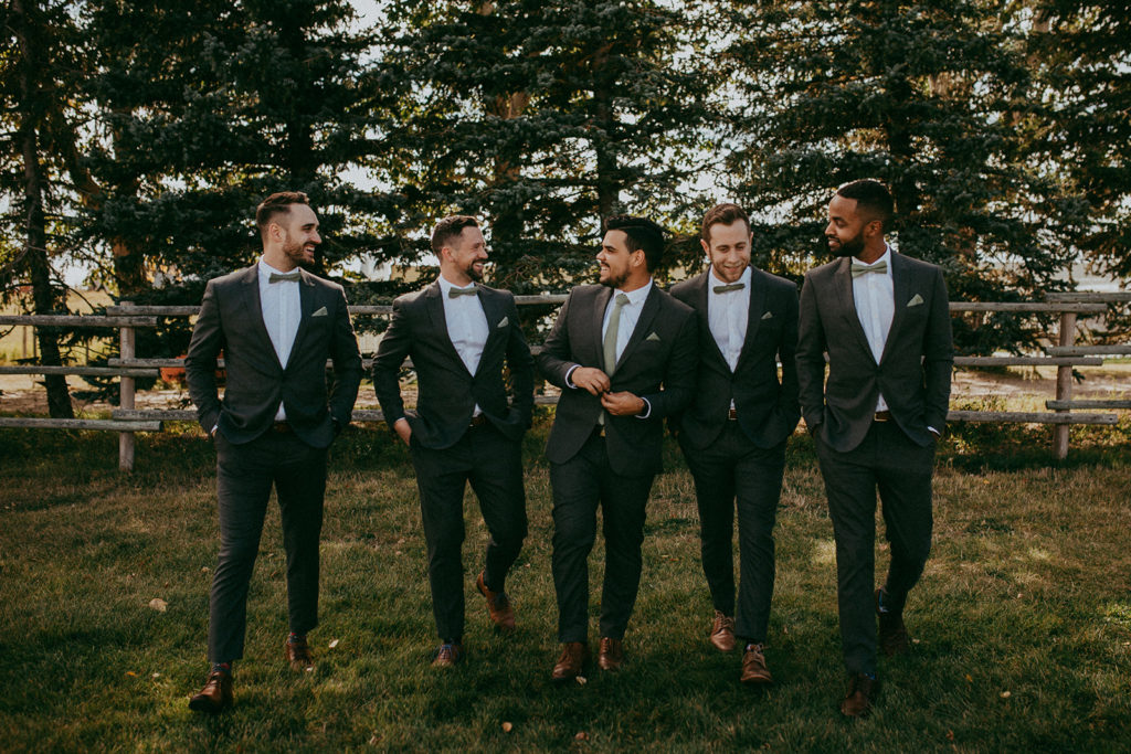 groomsmen walk casually in a grassy area with a fence and pine trees in the background