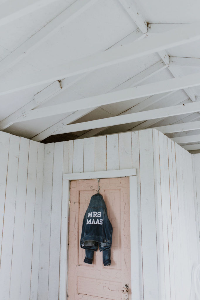 white cottage building with a blush pink door hangs a jean jacket with the brides new last name on the back