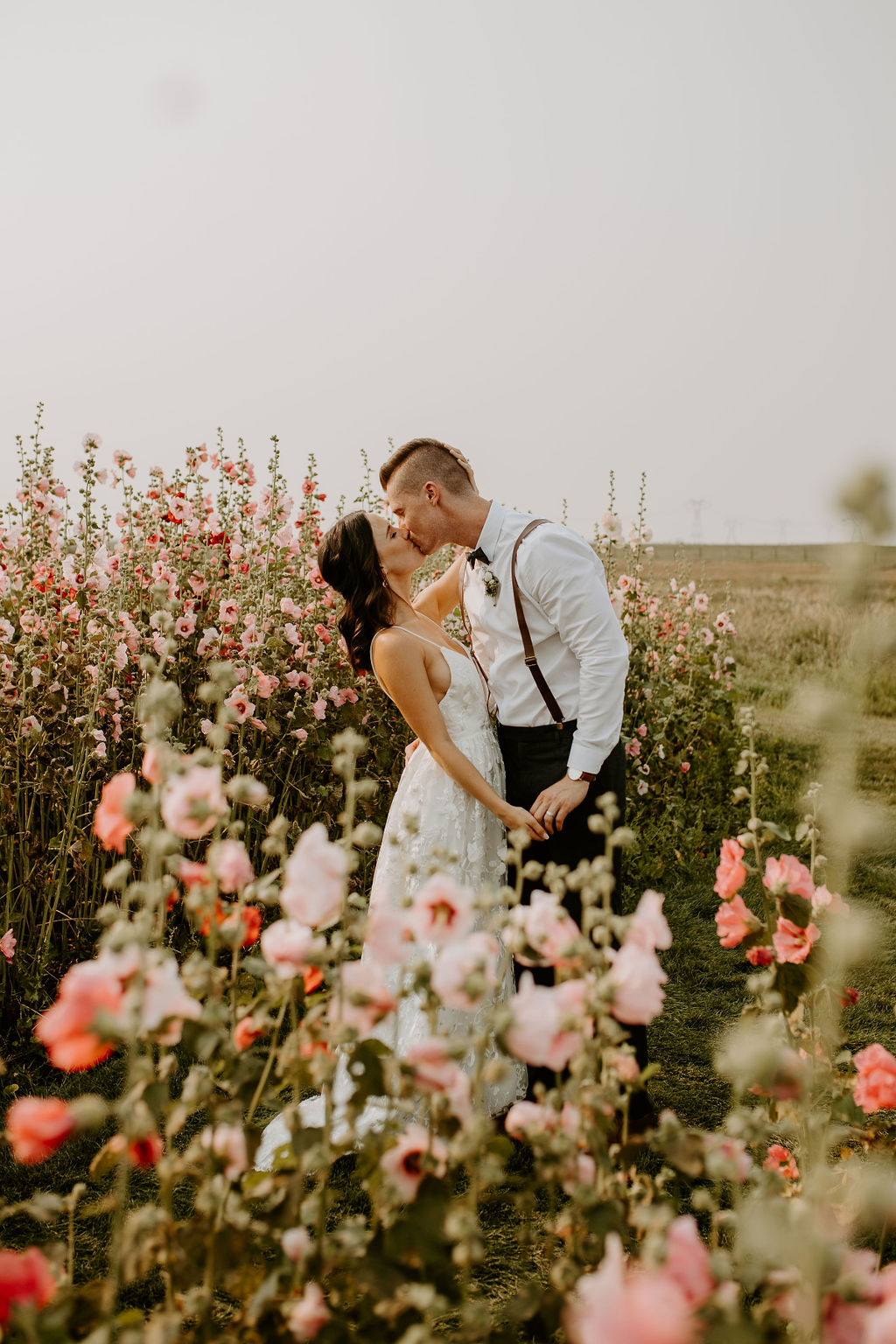 A bride and groom sharing a tender kiss in a field of pink hollyhocks, with a warm breeze gently caressing their faces.