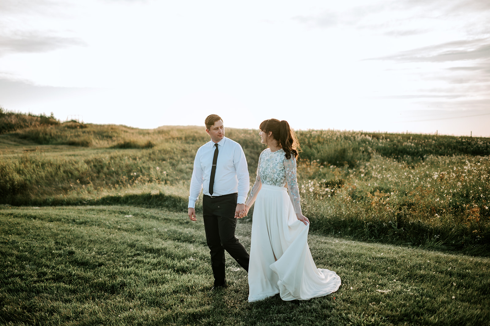 An artful day with a bride and groom walking through a grassy field at sunset.